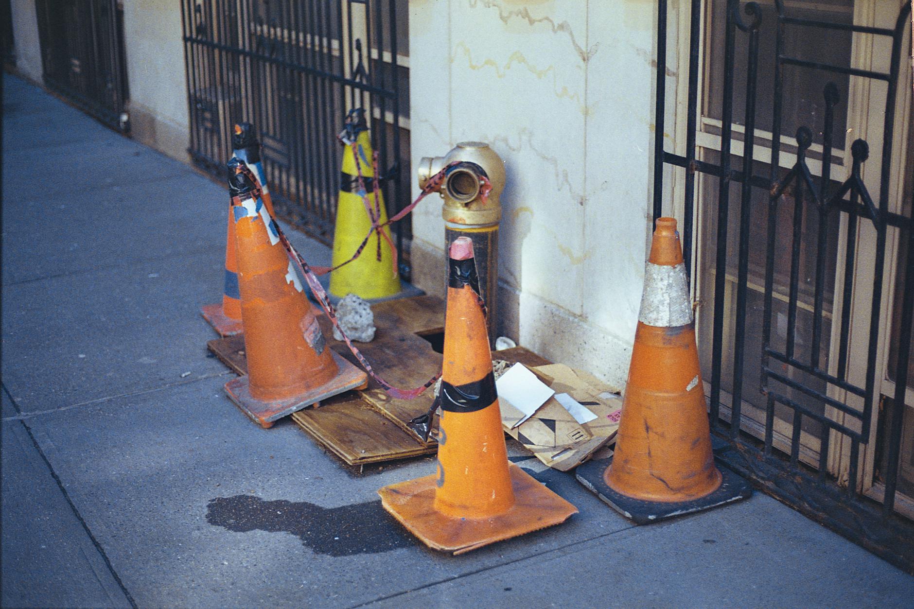 Sidewalk scene in NYC featuring traffic cones, a fire hydrant, and urban elements.