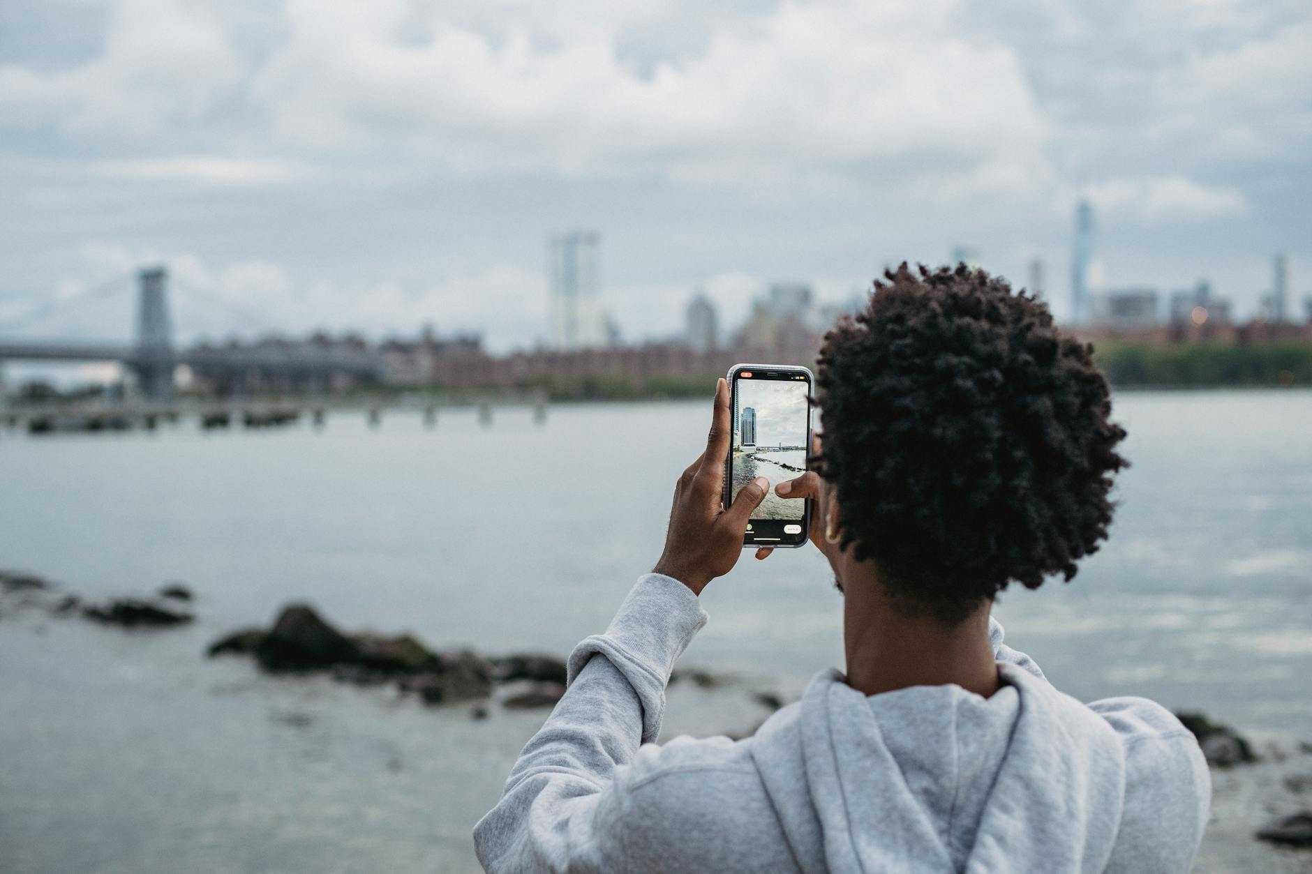 A person captures an urban skyline across the river using their smartphone, creating a serene outdoor moment.
